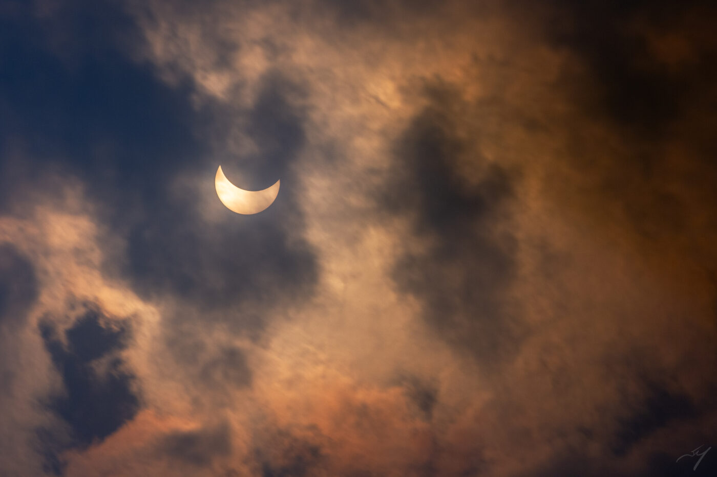 A partially-obscured sun peeks out behind orange-tinted clouds during a solar eclipse.
